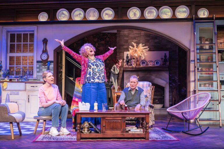 Dame Maureen Lipman as Allegra stands with her arms raised while Elizabeth Bower as Anna and John Middleton as Ronen sit in the cottage.