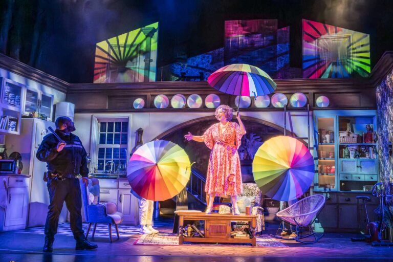 Dame Maureen Lipman as Allegra stands beneath rainbow umbrellas while Bailey Patrick as Officer Rogers watches during a colourful musical sequence.