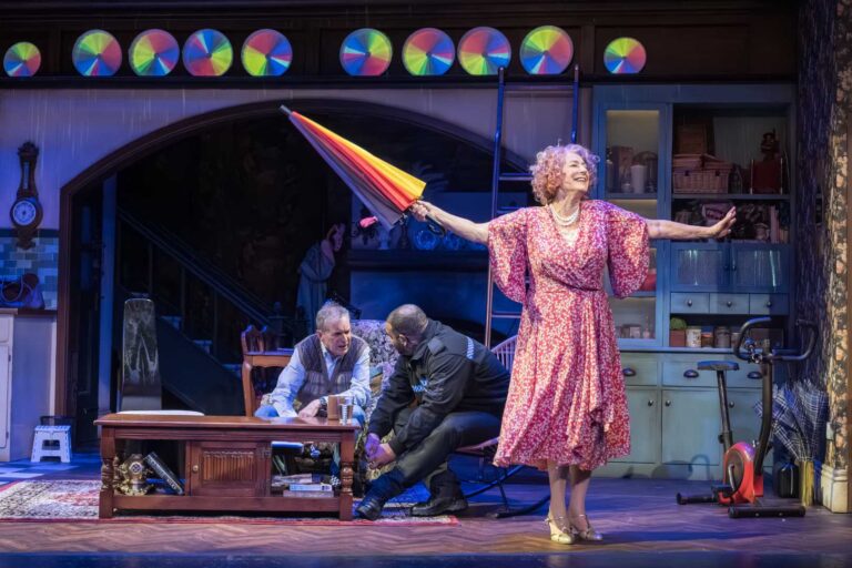 Dame Maureen Lipman as Allegra performs with a closed rainbow umbrella while John Middleton as Ronen and Bailey Patrick as Officer Rogers sit behind her.