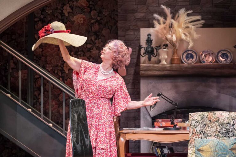 Dame Maureen Lipman as Allegra holding up a wide-brimmed hat in the cottage sitting room.